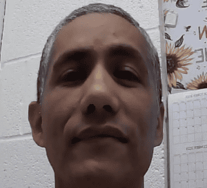 Close-up of an adult person with short gray hair looking at the camera, indoors against a light-colored wall; a calendar with sunflower images is visible in the background.