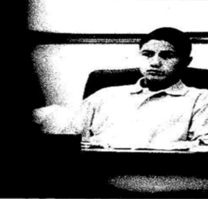 Black-and-white, high-contrast image of a young person sitting at a desk or table, facing forward with a neutral expression; the background is plain and the image appears grainy or stylized.